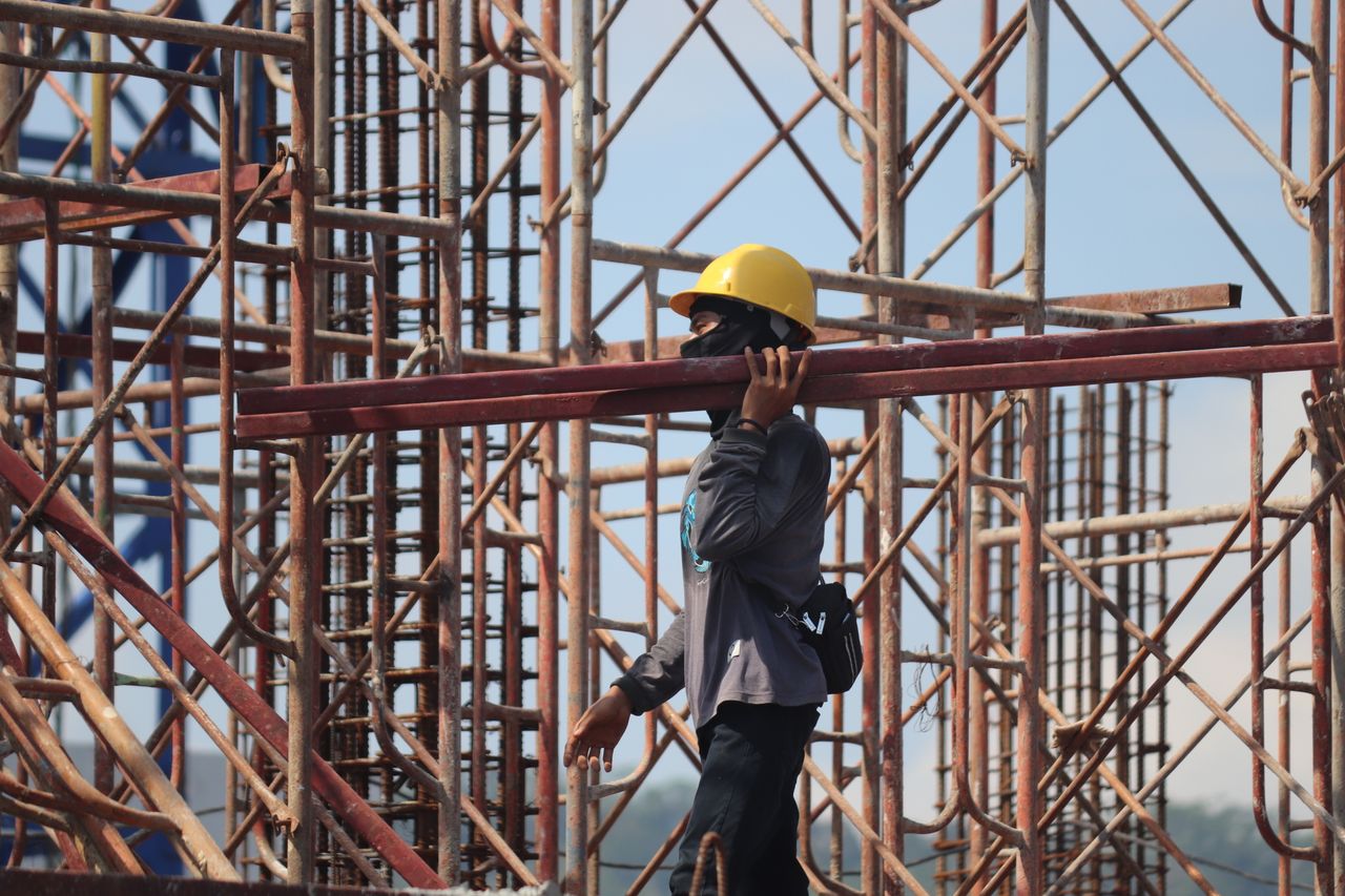 about-us-01 A construction worker with safety gear carrying a metal beam amidst scaffolding at a busy construction site.
